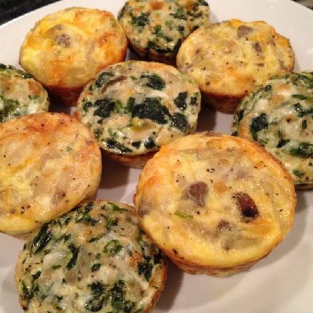 A plate of freshly baked, golden mini egg frittatas, each filled with a savory mixture that may include ingredients like cheese, vegetables, and meats.