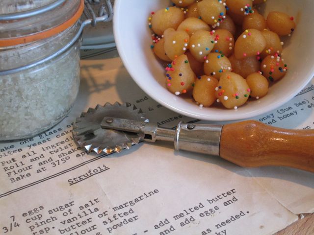 A bowl of round, traditional Italian struffoli beside a vintage pastry cutter, set on an open cookbook with a recipe visible, evoking a cozy, homemade baking atmosphere.