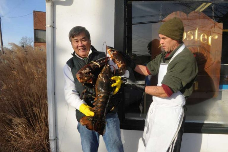Two men exchanging a large lobster outside The Lobster Bin in Greenwich, CT, with one man smiling, holding the lobster, and wearing gloves, while the other, in an apron, assists him