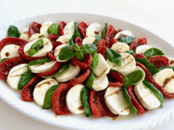Sliced tomato & mozzarella with fresh basil leaves are arranged on a platter and drizzled with balsamic glaze for a classic caprese presentation.