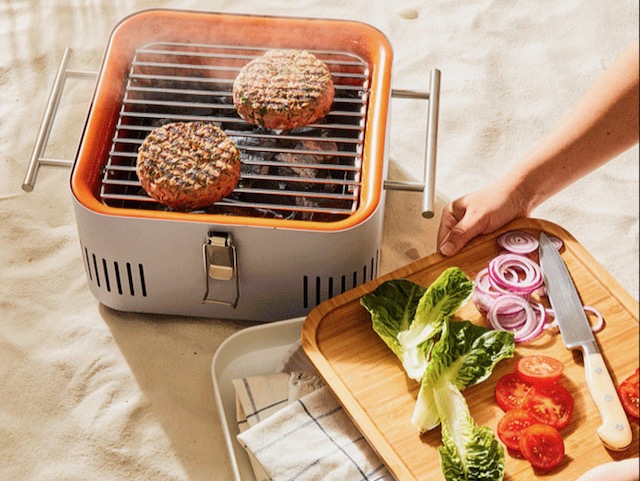 The Everdure CUBE Portable Charcoal Grill on the beach with two hamburgers grilling and vegetables shown in the chopping tray that also serves as a lid