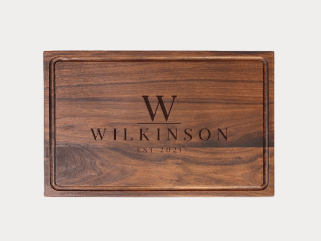 A personalized cutting board with the text 'WILKINSON EST 2021' and a large 'W' engraved above it. The wood boasts a rich, dark brown color with visible grain patterns, and the edges of the board are slightly raised.