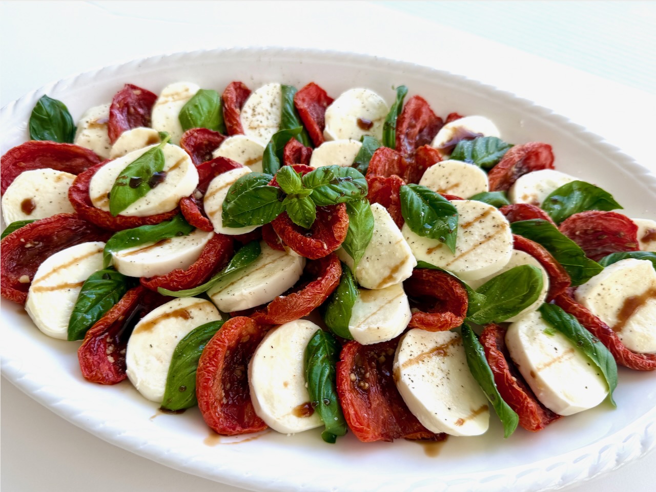 Large white platter with layers of sliced fresh Mozzarella, Slow roasted tomatoes, basil, and a drizzle of balsamic glaze. Caprese Salad.