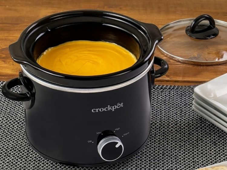 A black 2 quart slow cooker filled with creamy orange soup sits on a table beside a glass lid and stacked plates.