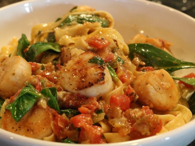 Pasta with seared scallops, spinach, and diced tomatoes in a white bowl. Pasta with seared scallops, spinach, and diced tomatoes in a white bowl.