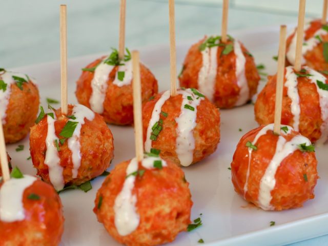 Slow cooker buffalo Chicken meatballs on a white platter with individual toothpicks and blue cheese dressing drizzled on top