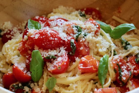 Spaghetti topped with cherry tomatoes, grated cheese, and fresh basil leaves in a wooden bowl.