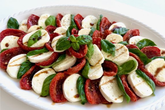Large white platter with layers of sliced fresh Mozzarella, Slow roasted tomatoes, basil, and a drizzle of balsamic glaze. Caprese Salad.
