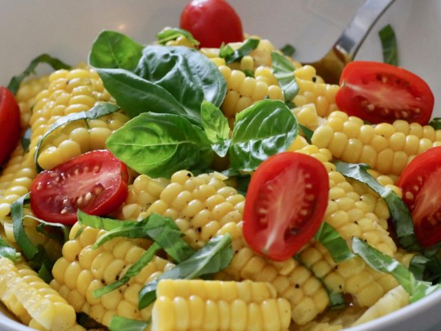Sliced corn salad with cherry tomatoes, fresh basil leaves, and black pepper in a white bowl.